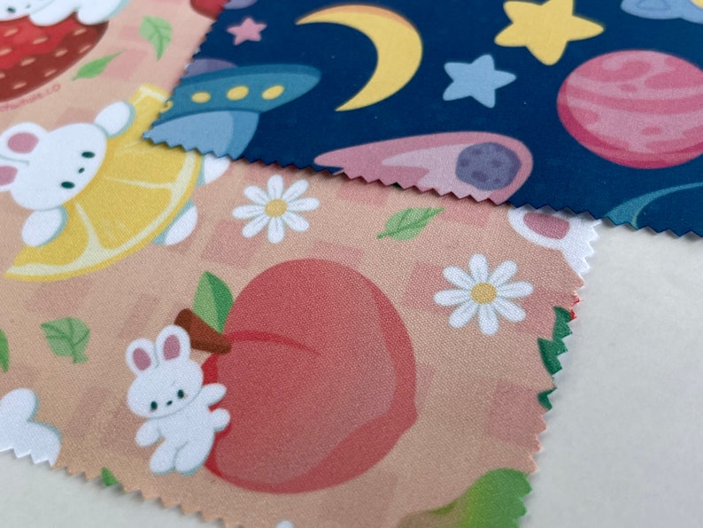 May include: Two fabric swatches with different patterns. The top swatch is navy blue with white stars, a yellow crescent moon, and a pink and purple planet. The bottom swatch is peach with white daisies, green leaves, and a pink peach with a white bunny.