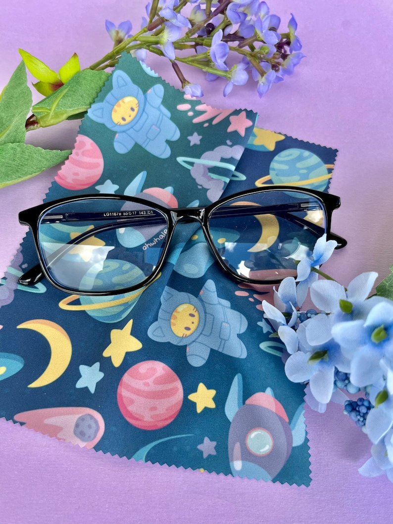 May include: Black framed glasses with blue lenses on a blue and black microfiber cleaning cloth with a space-themed pattern featuring planets, stars, rockets, and cats in astronaut suits. The text "ohwhale" is printed on the glasses.