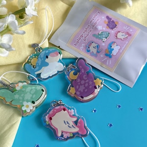 May include: A collection of holographic mystery phone charms featuring whimsical sea creature designs in various colors. The charms are attached to white lanyards and displayed on a blue surface with a packet that reads "holographic mystery phone charm".