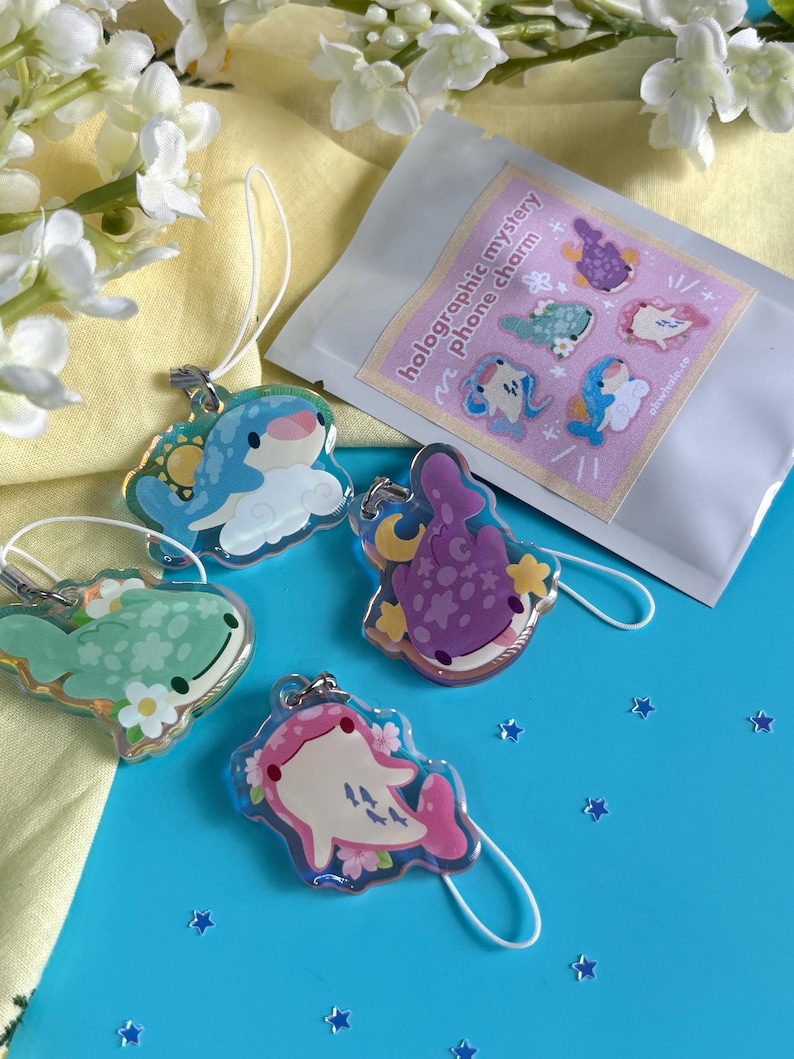 May include: A collection of holographic mystery phone charms featuring whimsical sea creature designs in various colors. The charms are attached to white lanyards and displayed on a blue surface with a packet that reads "holographic mystery phone charm".