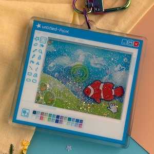 May include: A clear acrylic keychain featuring a whimsical design of a computer paint program with a clownfish illustration. The keychain has a rainbow-colored clasp and the text "untitled-Paint".