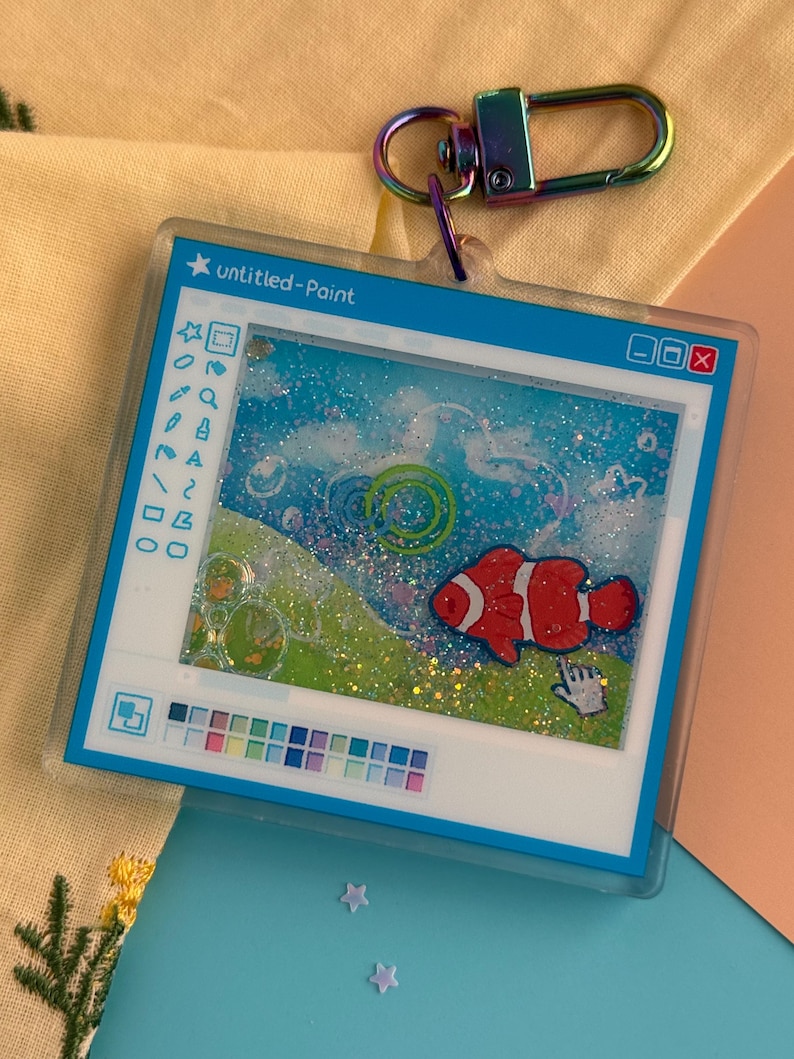 May include: A clear acrylic keychain featuring a whimsical design of a computer paint program with a clownfish illustration. The keychain has a rainbow-colored clasp and the text "untitled-Paint".