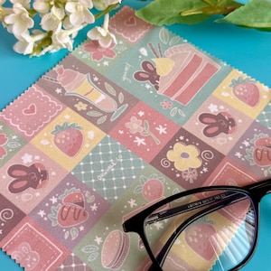 May include: A black-framed pair of eyeglasses rests on a patterned cleaning cloth. The cloth features a patchwork design with illustrations of desserts, strawberries, and bunnies. The background is a bright blue surface with white flowers.
