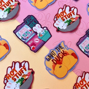 May include: A set of nine colorful stickers featuring cartoon bunnies and various phrases, including "What the Duck", "Hot Stuff", and "Gacha Addict". The stickers are on a pink and orange background.