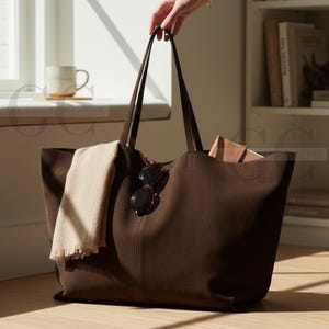 Op de afbeelding: Een grote, donkerbruine tote bag met lange handvatten wordt door een hand vastgehouden. De tas bevat een beige sjaal en een zonnebril. De tas staat op een houten vloer in de buurt van een raam en een boekenplank.