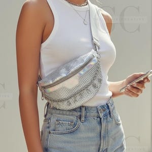 Op de afbeelding: Zilverkleurige heuptas met strass-ontwerp. De tas heeft een ritssluiting en een zilveren riem. Het woord "SASSY" is in strass-steentjes op de voorkant geschreven. De persoon draagt een witte tanktop en een spijkerbroekshort.