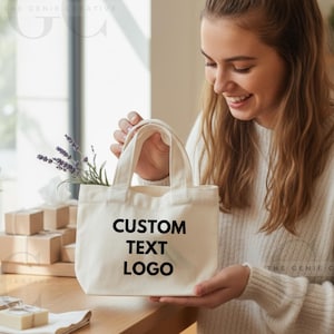 Peut inclure: Un sac fourre-tout crème avec le texte "CUSTOM TEXT LOGO" en noir. Le sac est tenu par une personne et contient de la lavande et des fleurs blanches. De petites boîtes marron sont en arrière-plan. Le sac a de courtes poignées.