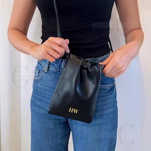 May include: Black leather drawstring bag with a long strap. The bag features gold-colored initials "HW" on the front. The bag is held by a person wearing a black top and blue jeans.