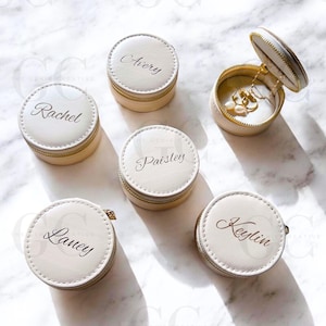 Könnte beinhalten: Cremefarbene, personalisierte Schmuck-Reisekoffer mit goldenen Reißverschlüssen. Jeder Koffer ist rund und trägt einen Namen in eleganter Schrift. Ein Koffer ist geöffnet und zeigt eine goldene Halskette und Ohrringe. Auf Marmoroberfläche angeordnet.