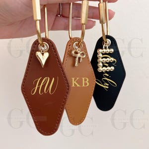 Könnte beinhalten: Drei personalisierte Schlüsselanhänger mit goldfarbener Hardware. Einer ist braun mit "HW", ein anderer ist hellbraun mit "KB" und der dritte ist schwarz mit "Emily". Jeder hat einen dekorativen Anhänger.