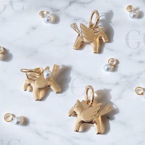 Puede incluir: Encantos dorados de Pegaso con detalles de perlas y pequeños aros dorados están dispersos sobre una superficie de mármol blanco. Los encantos presentan alas detalladas y están diseñados en forma de caballo estilizado. También hay pendientes adicionales de perlas y aros dorados.