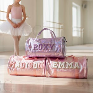 May include: Three iridescent pink duffel bags with names embroidered on them. The top bag says "ROXY" in purple glitter letters. The bottom bags say "MADISON" and "EMMA" in white and pink letters. A young girl in a ballet outfit is in the background.