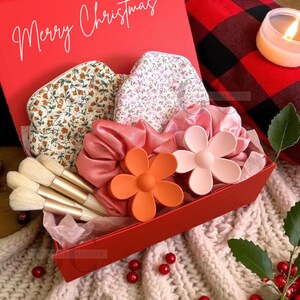 以下が含まれることがあります： 白い筆記体で「Merry Christmas」と書かれた赤いギフトボックス。中には花柄のシュシュ、花の形をしたヘアクリップ、メイクブラシ、サテンのシュシュが入っています。ボックスは白いニットの表面にあります。