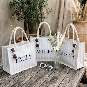 May include: Three light blue and white striped canvas tote bags with sturdy handles. Each bag features a personalized name in dark blue text: EMILY, ASHLEY, and JESSICA. Each bag is decorated with a pair of white heart-shaped sunglasses.