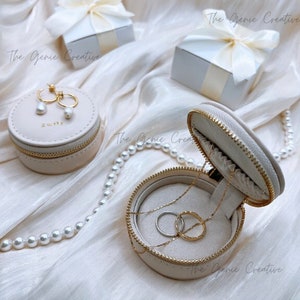 May include: A beige round jewelry box with a zipper closure. The box is open and contains a gold necklace and two rings. The box is sitting on a white surface with a pearl necklace.