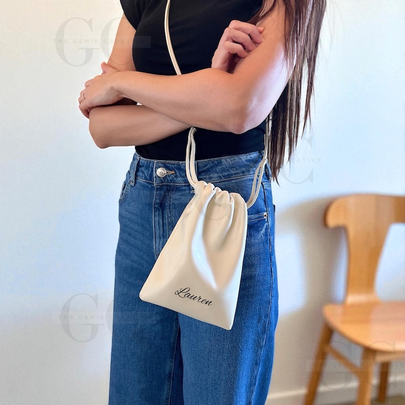 May include: Cream-colored drawstring bag with the name "Lauren" printed in black script. The bag is worn crossbody style with a thin, matching strap. The person is wearing a black top and blue jeans.