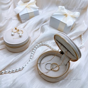May include: Two beige zippered jewelry boxes with gold accents. One box is open and contains a gold necklace and two gold rings. The other box is closed and contains a pair of pearl earrings. The boxes are on a white fabric background with a pearl necklace.