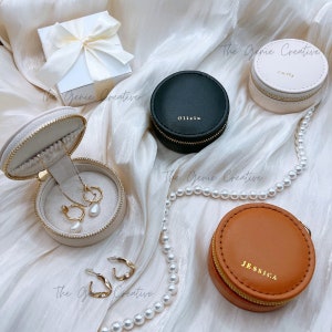 May include: Four round jewelry boxes with zippers. The boxes are beige, black, brown, and white. The boxes have gold lettering with the names "Olivia", "Sally", "Jessica", and "The Genie Creative".