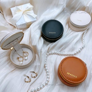 May include: Three round jewelry boxes with zippers. The boxes are beige, black, and brown. The beige box has a pearl necklace and earrings inside. The black box has the name "Olivia" on it. The brown box has the name "Jessica" on it.