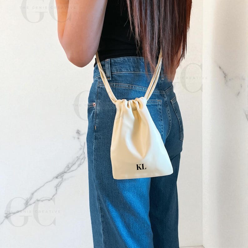 May include: A cream-colored drawstring bag with the initials "KL" printed in black. The bag is suspended by a thin, cream-colored cord. The bag is hanging in front of a person wearing blue jeans and a black top.