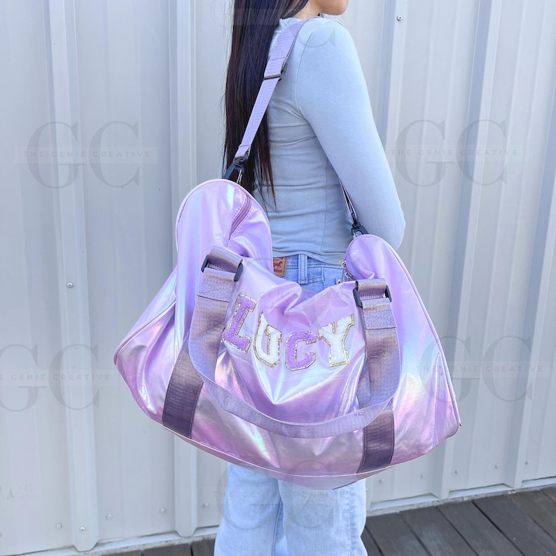 May include: A purple iridescent duffel bag with a black strap and the name "Lucy" in white and gold letters.
