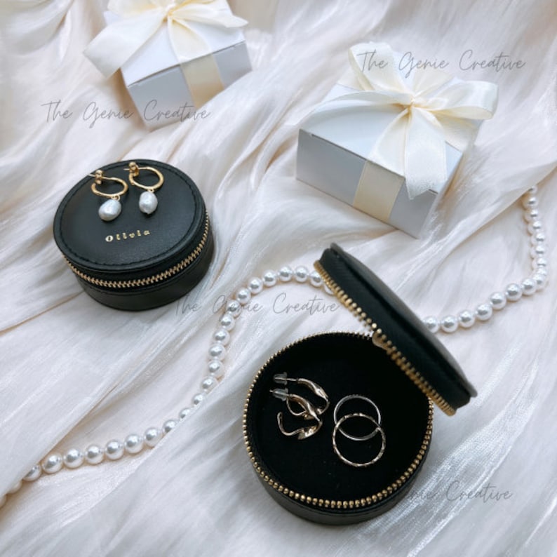 May include: Two black round zippered jewelry boxes with gold zippers. The smaller box has a white label with the word "Olivia" on it. The larger box is open and has gold earrings and rings inside. The boxes are on a white fabric background with a pearl necklace.