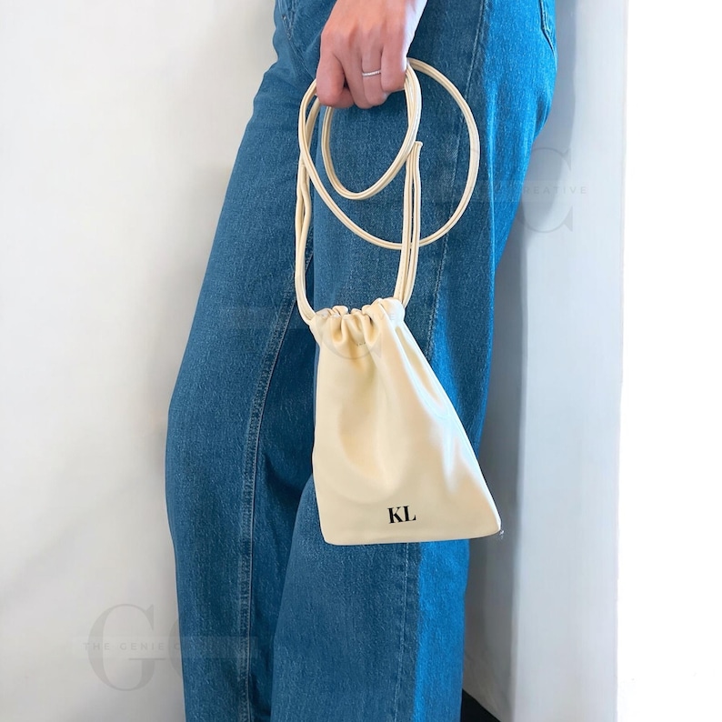 May include: A cream-colored drawstring bag with a long, thin strap. The bag has the initials "KL" printed in black on the front. The bag is held against a background of blue denim jeans.