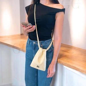 May include: A cream-colored drawstring phone bag with the initials "KL" printed in black. The bag is suspended from a thin, matching strap. The person is wearing a black top and blue jeans.