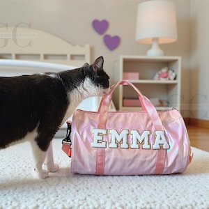 以下が含まれることがあります： 白と金色の文字で「EMMA」と書かれたピンク色のダッフルバッグ。バッグにはピンク色のハンドルとショルダーストラップが付いています。白黒の猫がバッグを嗅いでいます。バッグは白いラグの上にあります。