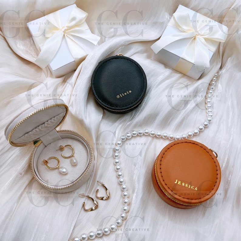 May include: Three round jewelry boxes with different colored leather exteriors. The boxes are on a white fabric background with a pearl necklace. The boxes are labeled "Olivia", "Jessica", and "The Genie Creative".