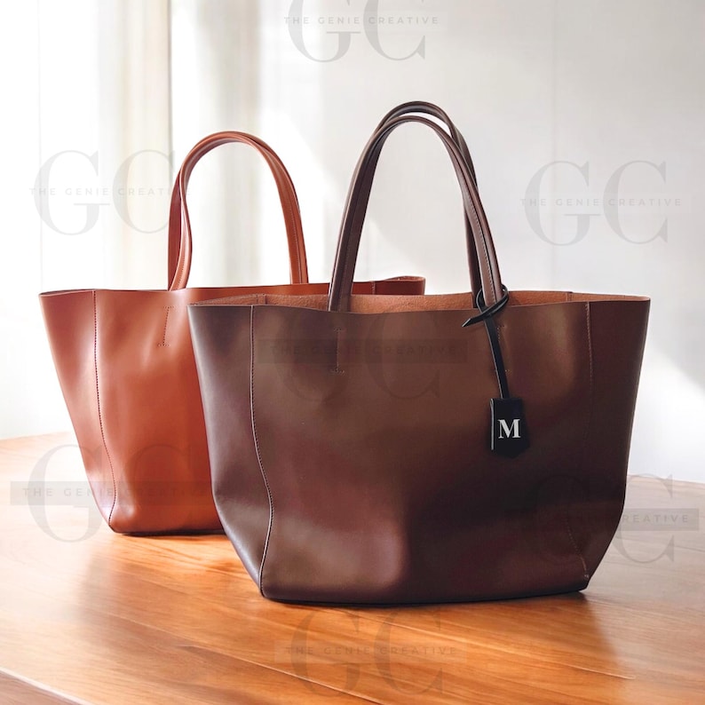 May include: Two leather tote bags, one brown and one dark brown. The dark brown bag has a black leather tag with the letter "M" on it.