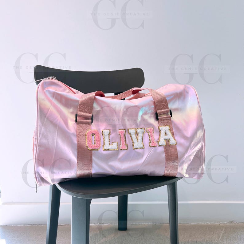 May include: A pink iridescent duffel bag with a white and gold name patch that reads "OLIVIA".