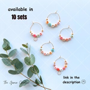 May include: Set of five gold-tone wine glass charms with colorful beads spelling out names. The charms are decorated with small flower accents. The text "available in 10 sets" is at the top of the image. The text "link in the description" is at the bottom of the image.