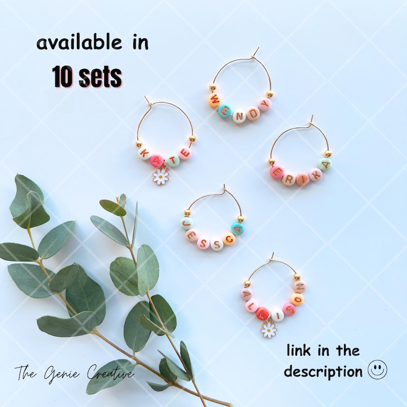 May include: Set of five gold-tone wine glass charms with colorful beads spelling out names. The charms are decorated with small flower accents. The text "available in 10 sets" is at the top of the image. The text "link in the description" is at the bottom of the image.