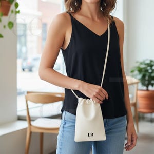 May include: Cream-colored crossbody bag with a drawstring closure and a long, thin strap. The bag has the initials "LB" printed in black on the front. The person in the image is wearing a black tank top and blue jeans.