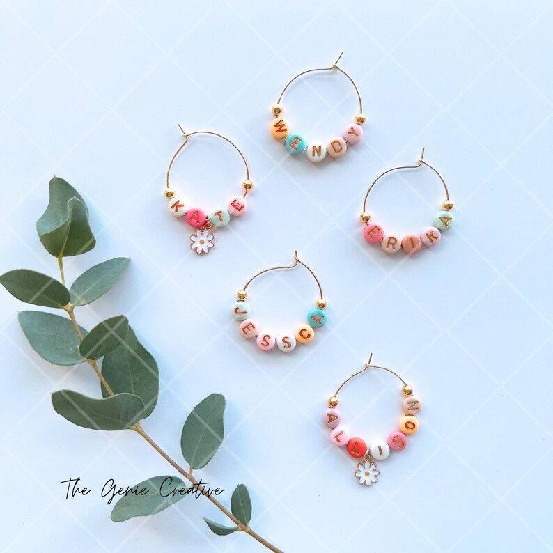 May include: Five gold-colored wine glass charms with colorful beads spelling out the names "Wendy", "Katie", "Erika", "Jessica", and "Allison". Each charm has a small white flower charm attached.