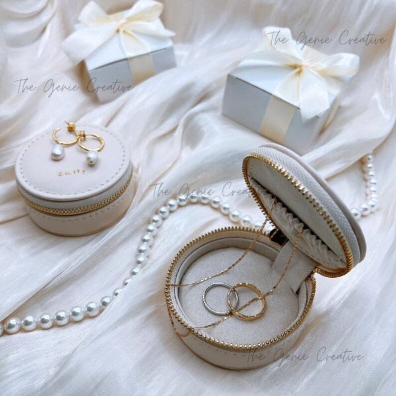 May include: A beige round jewelry box with a zipper closure. The box is open and contains a gold necklace and two rings. The box is sitting on a white surface with a pearl necklace.