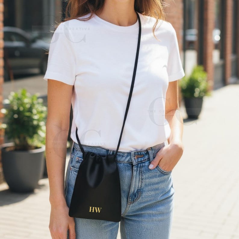 May include: Black drawstring crossbody bag with gold-colored initials "HW". The bag is worn over the shoulder with a thin black strap. The person is wearing a white t-shirt and blue jeans.