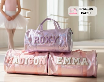 Op de afbeelding: Drie cilindervormige sporttassen met een glanzende, iriserende afwerking. De bovenste tas heeft de naam ROXY in paarse glitterletters. De onderste twee tassen hebben de namen MADISON en EMMA in witte en gouden glitterletters.