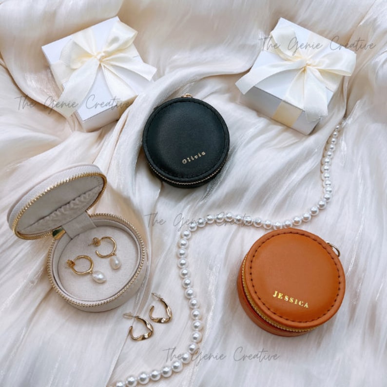 May include: Three round jewelry boxes with zippers. The boxes are beige, black, and brown. The beige box has gold earrings inside. The black box has the name "Olivia" on it. The brown box has the name "Jessica" on it. A string of white pearls is in the background.