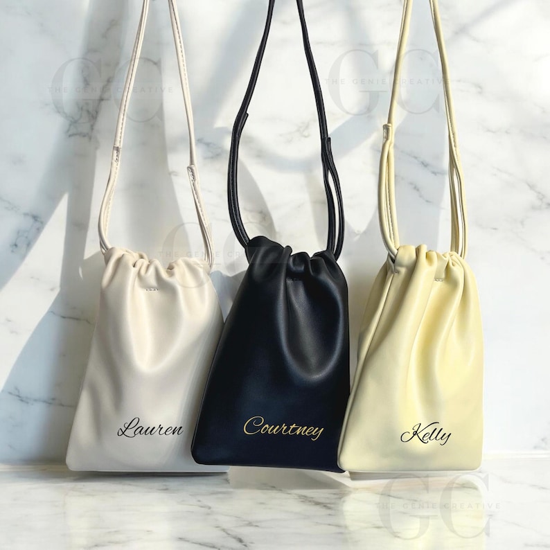 May include: Three personalized drawstring bags in cream, black, and pale yellow. Each bag has a long strap and a name printed in a cursive font. The cream bag is labeled "Lauren", the black bag is labeled "Courtney", and the yellow bag is labeled "Kelly".