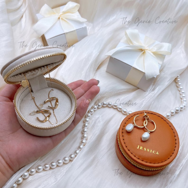 May include: Two round jewelry boxes, one beige and one brown. The beige box is open and contains a gold necklace and earrings. The brown box is closed and has the word "JESSICA" printed on the lid. Both boxes have a zipper closure.