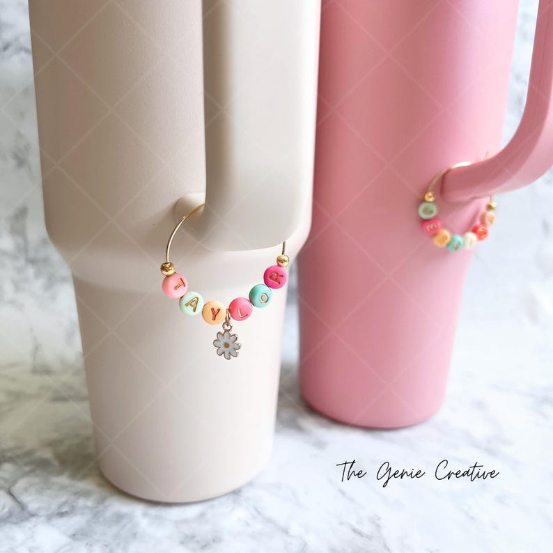 May include: Two pink and white ceramic mugs with gold-colored metal rings attached to the handles. The rings are decorated with colorful beads that spell out the names "TAYLOR" and "BROOK." Each ring has a small white flower charm attached.