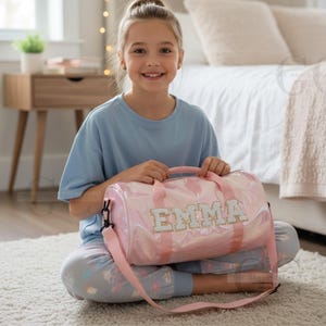 以下が含まれることがあります： キラキラした文字で「EMMA」と書かれたピンク色のダッフルバッグ。バッグにはショルダーストラップとハンドルが付いています。バッグは光沢のある素材で作られています。バッグは白いカーペットの上にあります。