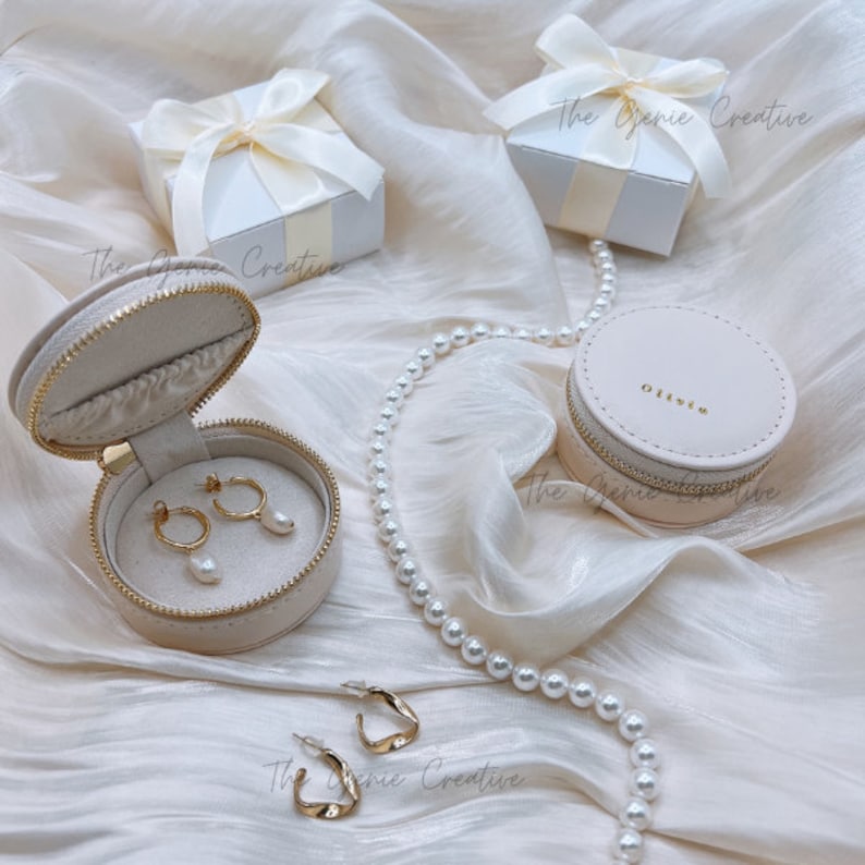 May include: Two round beige jewelry boxes with zippers, one with a pearl necklace and a pair of pearl earrings inside, the other with a pair of gold hoop earrings inside. The boxes are on a white satin fabric background. The text "The Genie Creative" is visible in the image.