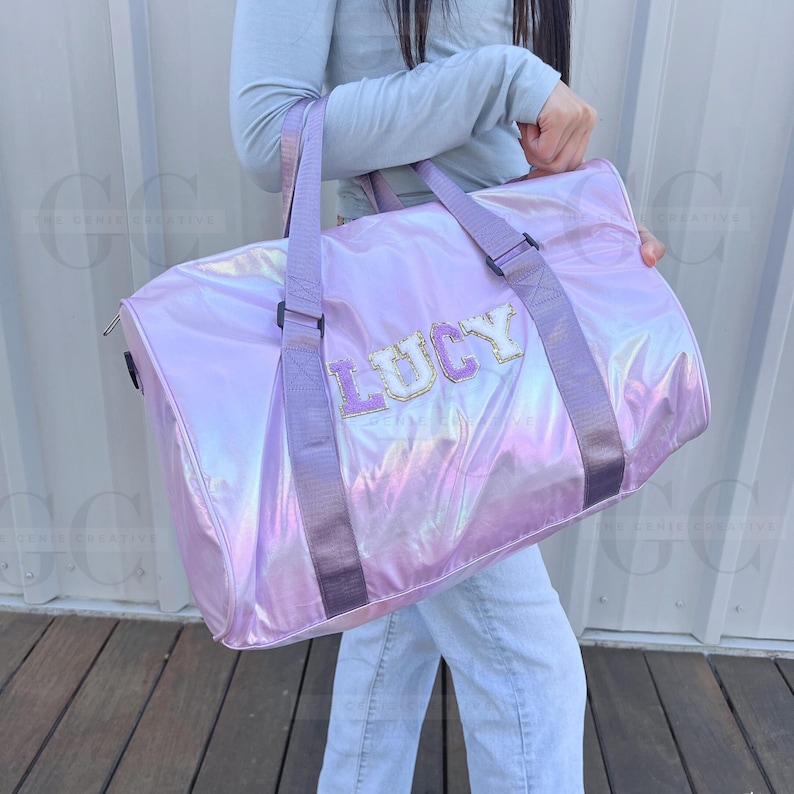 May include: A light purple iridescent duffel bag with black straps and the name "LUCY" in white letters with a pink outline.