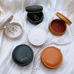 May include: Six round, zippered jewelry boxes in various colors, including black, white, brown, and tan. Each box has a different name on it, including Olivia, Emily, and Jessica. The boxes are on a white surface with a pearl necklace.
