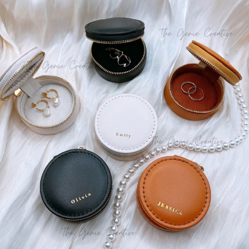 May include: Six round, zippered jewelry boxes in various colors, including black, white, brown, and tan. Each box has a different name on it, including Olivia, Emily, and Jessica. The boxes are on a white surface with a pearl necklace.