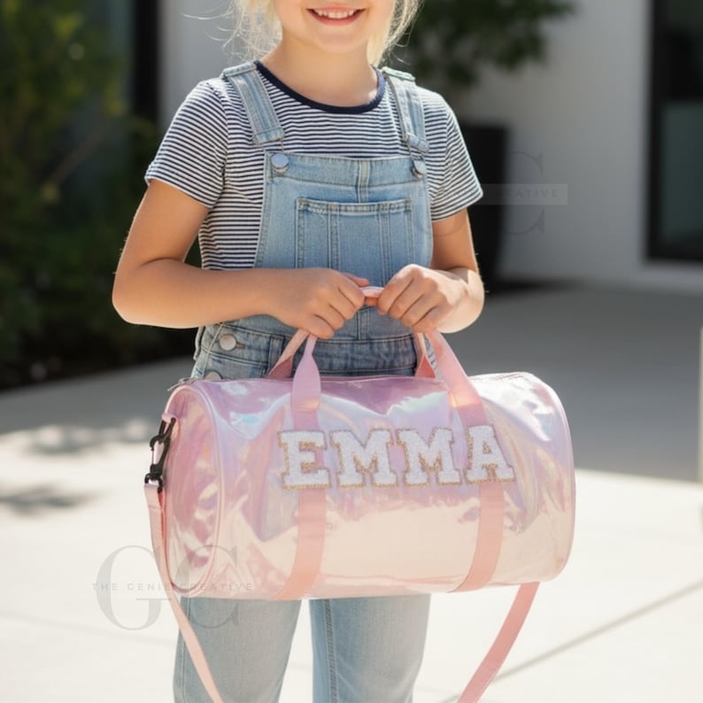 以下が含まれることがあります： 白いブロック文字で「EMMA」という名前が入ったピンク色のイリデセントダッフルバッグ。バッグにはピンク色のハンドルとショルダーストラップが付いています。バッグを持っている人は、デニムのオーバーオールとストライプのシャツを着ています。