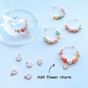May include: Five gold-colored wine glass charms with colorful beads and letters spelling out names.  Each charm has a small white daisy charm attached.  The text "Add flower charm" is written below the charms.